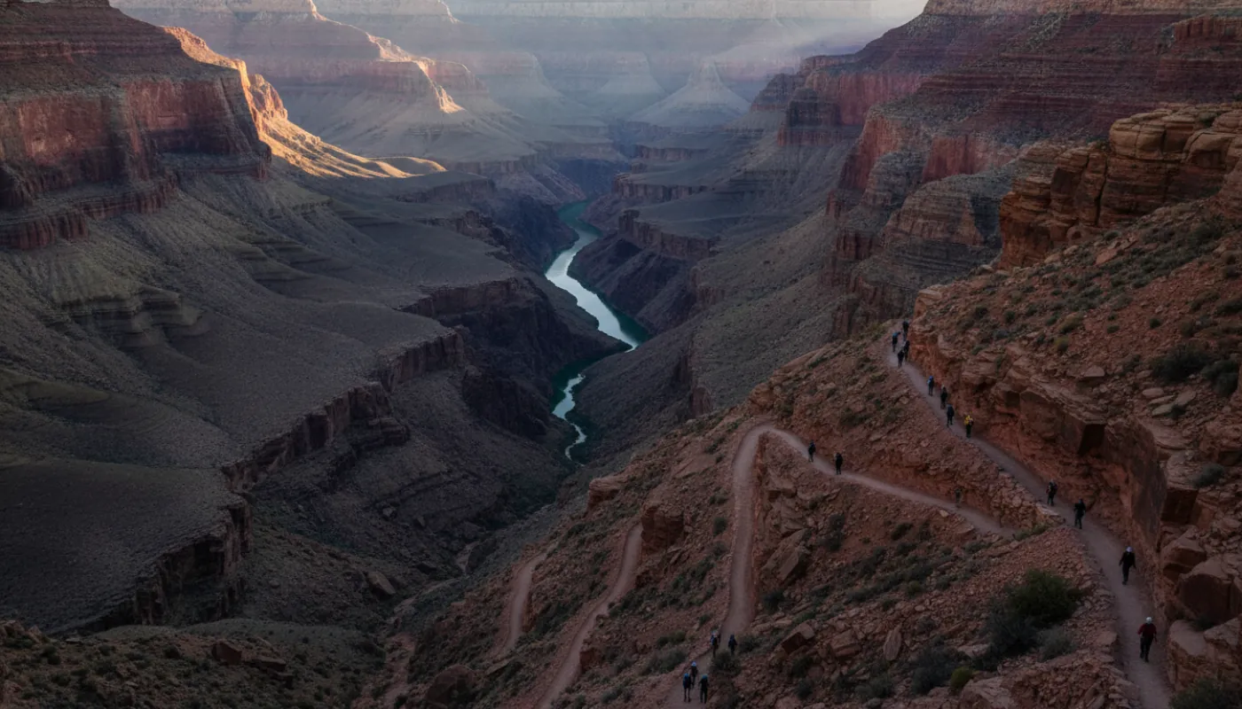A line of hikers descending a switchbacking corridor trail into the Grand Canyon at dawn, red rock walls and the distant Colorado River far below, cool early light